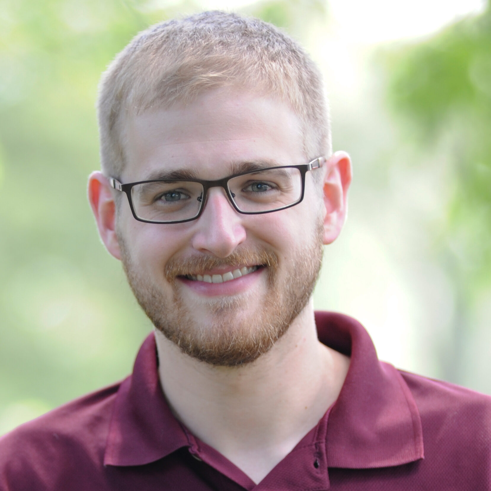 Young man in a maroon polo, with short blond hair and black glasses.