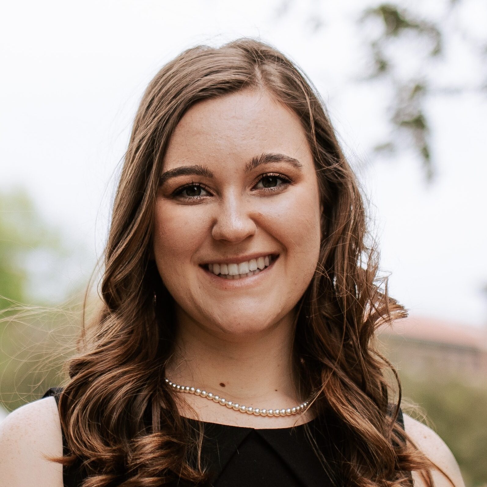 Headshot of a young woman wearing a black dress and a string of pearls. She has brown hair that has been curled.