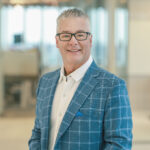 headshot of Daryl Knecht - light hair, black glasses, white shirt and blue checked jacket standing in a office area.