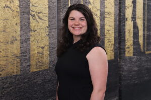 Head-shot of a brunette woman in a black top standing in front of a gold and black background.
