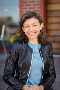 Women with dark medium length hair in a light blue shirt with a leather dark jacket.  Head shot with brick and cedar siding in the background.