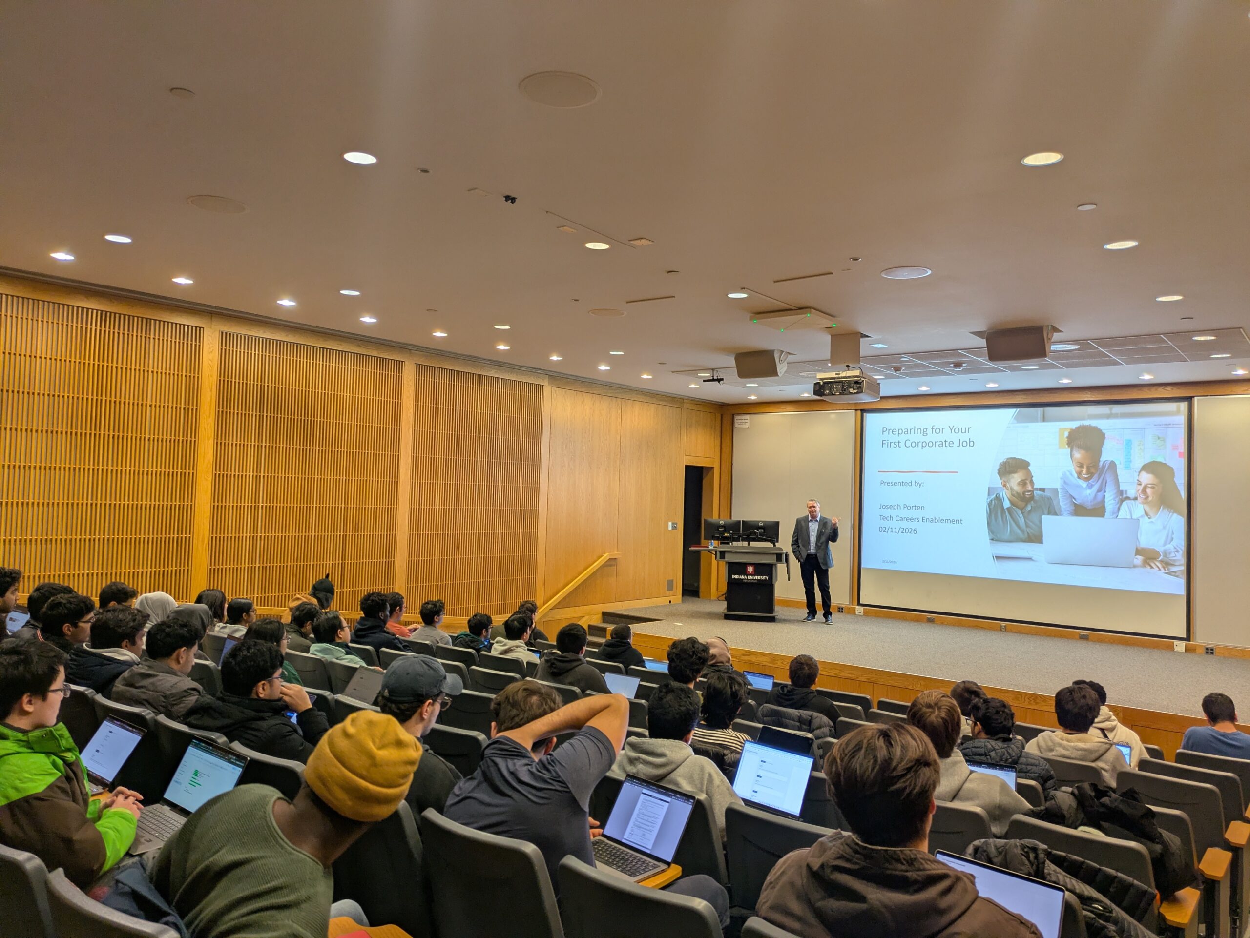 Students seated in an auditorium. There is a speaker on an elevated stage.