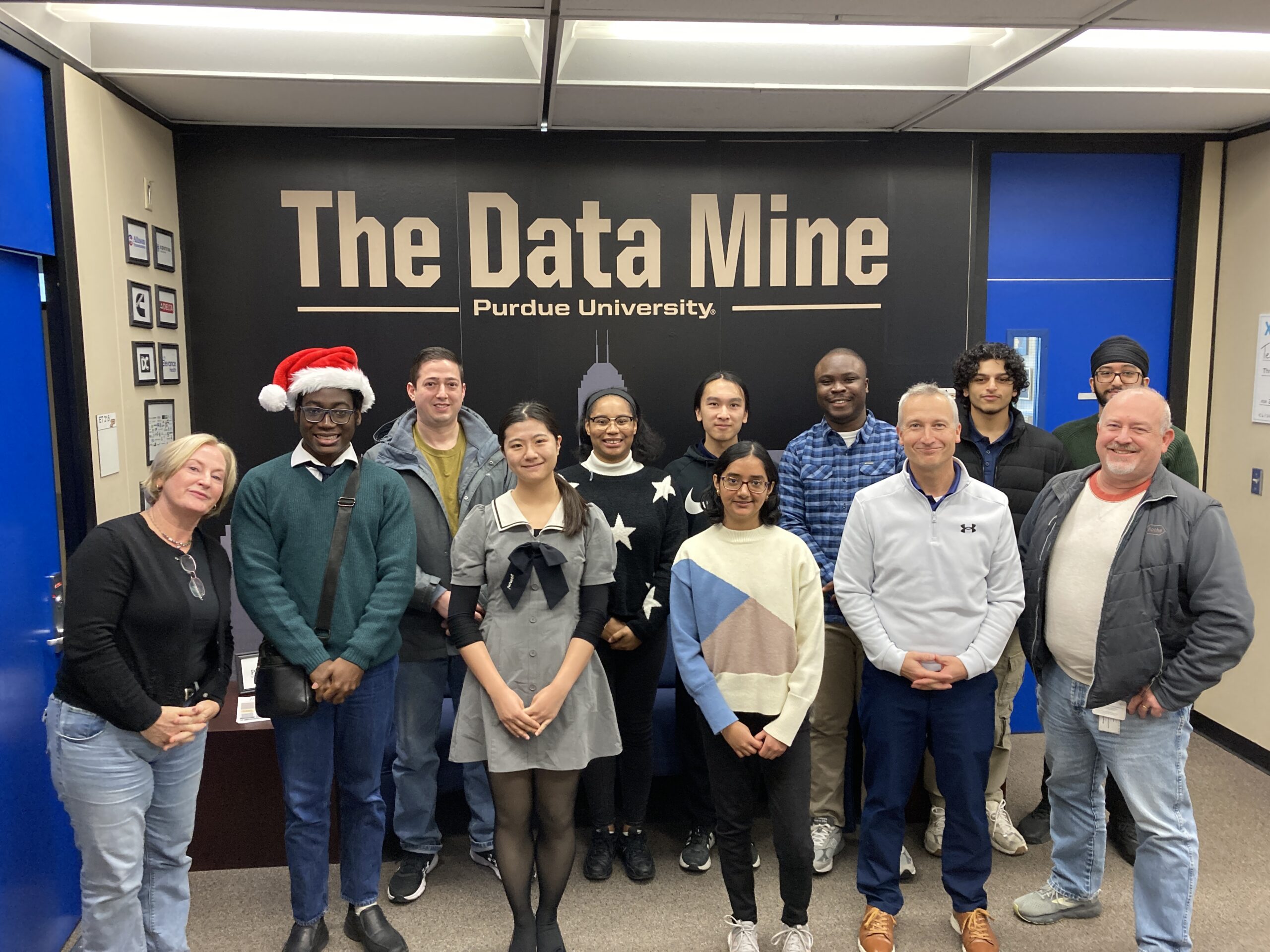 A group of students stand with their mentors in front of a large black sign that says "The Data Mine".